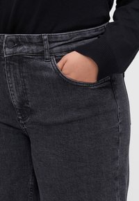 Černé džíny z denimu s úzkým střihem, vybavené přední kapsou, poutky na opasek a kontrastním prošíváním. Materiál působí pevně a má jemnou texturu.