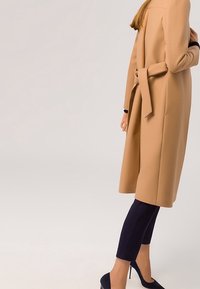 Abrigo largo beige con un acabado texturizado, corte ajustado, cinturón atado en la cintura y aberturas laterales. Combinado con pantalones cortos azul marino y tacones negros.