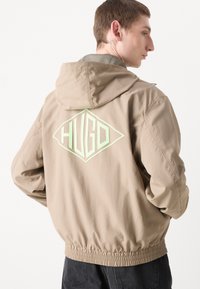 Beige hettegenser med elastiske cuff, med et diamantformet lys grønt "HUGO"-logo på ryggen. Glatt finish og uformelt design.