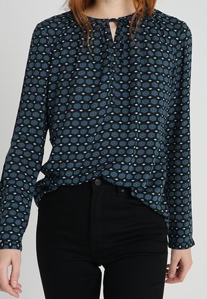 Femme portant un chemisier à manches longues à motif avec un col noué et un pantalon noir, montrée des épaules aux hanches.