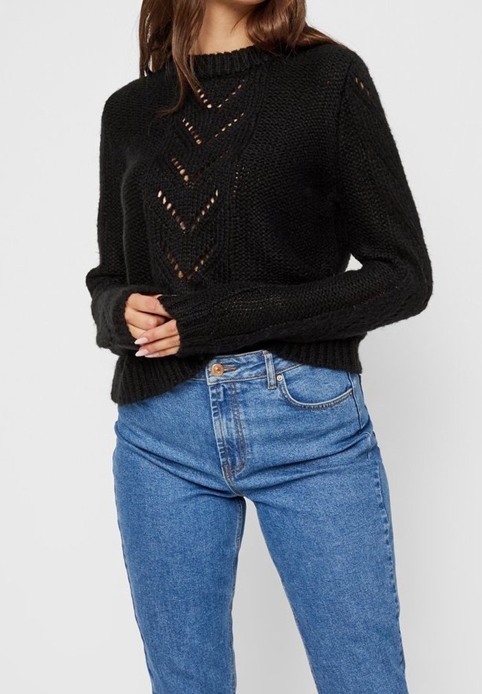 Femme portant un pull en maille noire avec des motifs œillets en chevron et un jean bleu taille haute, debout, les mains près de la taille.