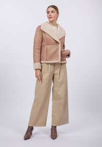 Faux semsketjakk i lys brun med en kremfarget saueskinnskrage og mansjetter, med en enkelt knappelukking og strukturert design. Kombinert med vide beige bukser.
