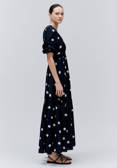 Kobieta w czarnej długiej sukience w grochy z bufkami na rękawach, stojąca bokiem, nosząca czarne sandały na płaskim obcasie na paski oraz złote pierścionki.