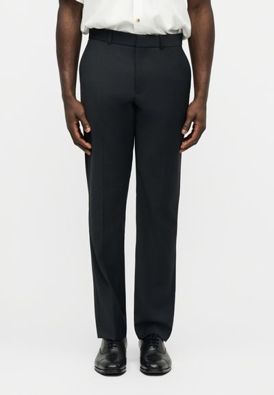 Homme portant un pantalon noir sur mesure, des chaussures habillées noires et une chemise blanche rentrée, debout devant un fond blanc uni.