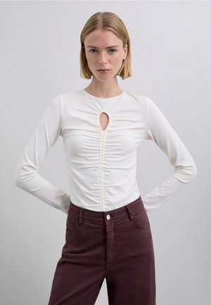 Haut blanc à manches longues avec une découpe en goutte d'eau et un détail froncé à l'avant, associé à un pantalon bordeaux taille haute, affichant une texture lisse.
