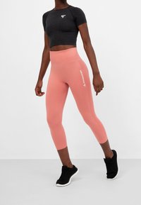 Top corto nero a maniche corte con logo bianco, abbinato a leggings pesco a vita media con testo "SQUATPROOF", indossato con sneakers nere.
