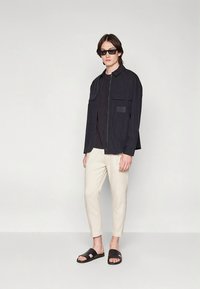 Černá zipová bunda s dvěma předními kapsami, nošená přes černé tričko. Světlé zúžené kalhoty a černé sandály. Stylové sluneční brýle doladí vzhled.