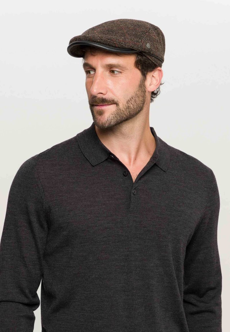 Brązowa czapka typu flat cap w wzory, ze skórzanym wizjerem, noszona przez mężczyznę w ciemnej, długosleeve'owej koszulce polo, prezentująca teksturowany materiał przypominający wełnę.