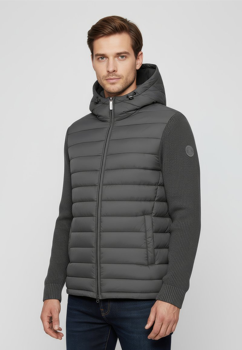 Mann trägt eine graue wattierte Kapuzenjacke mit Strickärmeln und Reißverschluss vorne, steht vor einem schlichten hellen Hintergrund.