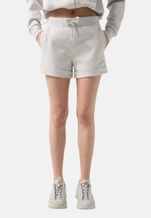 Persoon draagt lichtbeige shorts met trekkoord en opgerolde zomen, en bijpassende sneakers, handen in de zakken, tegen een effen achtergrond.