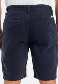 Tummansinisiä shortsit, jotka on valmistettu puuvillasta ja joissa on sileä pinta. Shortsien takana on kaksi taskua, joissa on napit, ja oikealla puolella on logopatch.