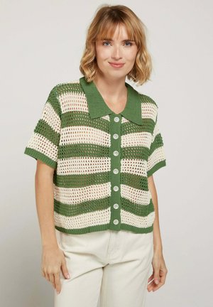 Femme aux cheveux blonds courts portant une chemise crochetée à rayures vertes et crème avec boutons et un pantalon de couleur claire, souriant doucement.