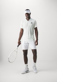 Polo shirt de tennis blanc avec des accents noirs, associé à un short blanc et des baskets. Tenant une raquette de tennis, portant une visière blanche.