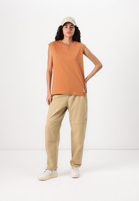 Haut sans manches orange et pantalon beige avec des poches cargo. La personne porte des chaussures blanches et une casquette de couleur claire. Design simple et fonctionnel.