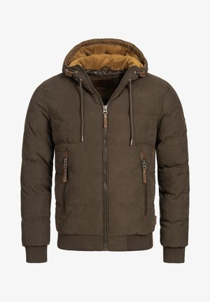 Homme portant une veste matelassée marron avec une capuche bordée de fourrure, une chemise blanche, un jean gris clair et des baskets blanches, debout avec la main dans la poche.