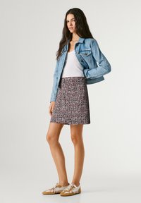 Młoda kobieta stojąca, ubrana w jasnoniebieską kurtkę dżinsową, biały top na ramiączkach, spódnicę w wzory oraz beżowo-białe buty sportowe na jednolitym tle.