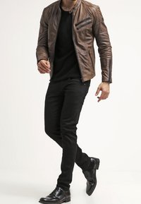 Hnědá kožená bunda se zipy, černý svetr, slim černé kalhoty a černé lesklé boty. Celkový vzhled je elegantní s přiléhavým střihem.