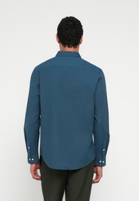 Žalsvai mėlynas ilgomis rankovėmis marškiniai su užsegamu apykakliu, tekstūruota medžiaga, apvalia apačia ir užsegamais rankogaliais, dėvimi su tamsiomis kelnėmis, matomi iš nugaros.