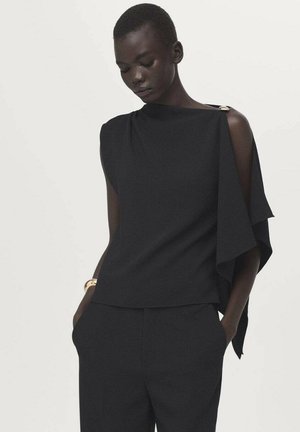 Ženska s kratkimi lasmi, oblečena v črno brezrokavno majico z drapiranim detajlom na rami in ujemajoče črne hlače, z rokami v žepih.
