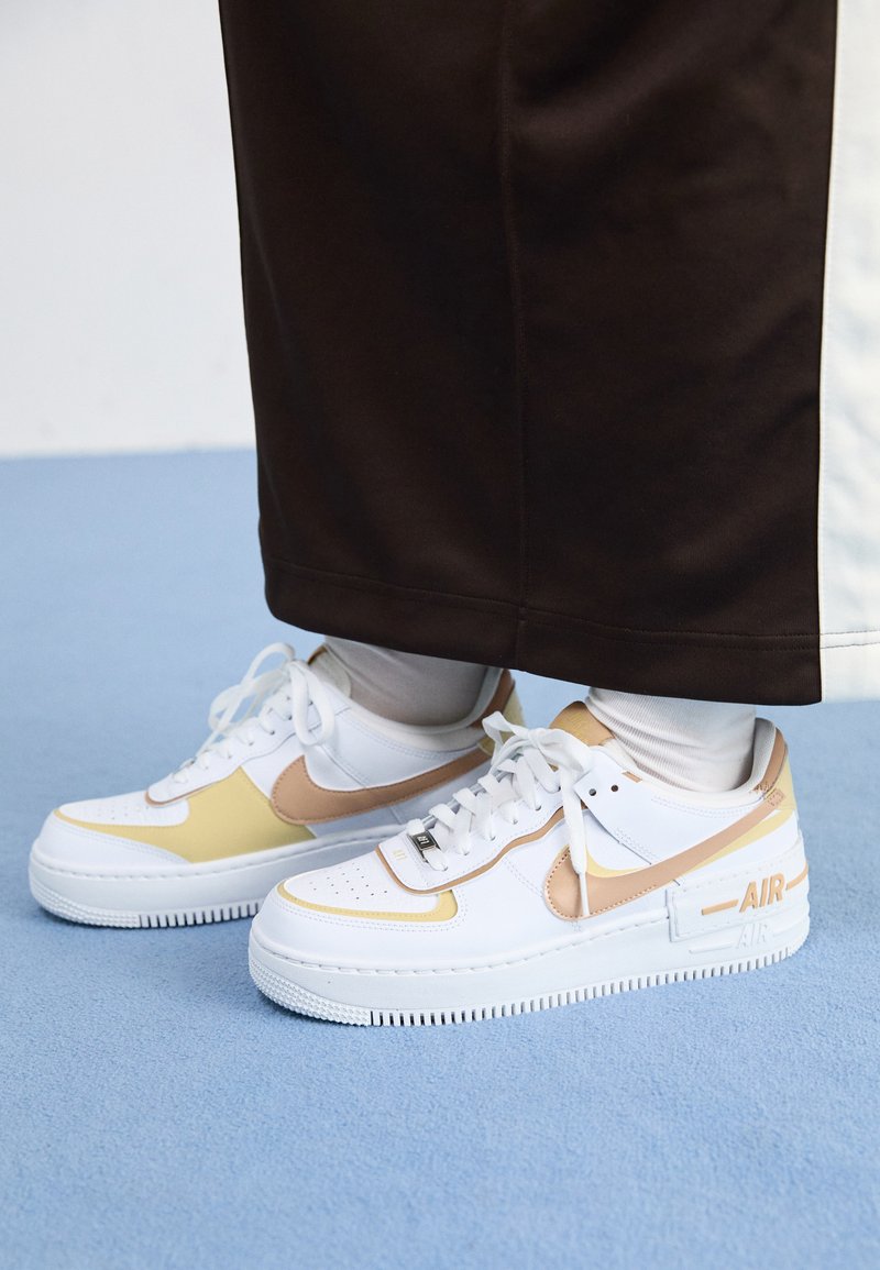 Zapatillas de cuero blancas con acentos dorados, que cuentan con una puntera perforada, cordones planos y una suela de goma gruesa con el logotipo "AIR" en el lateral.