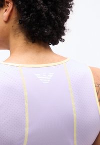 Άτομο με σγουρά μαλλιά που φοράει αμάνικη μπλούζα ανοιχτού μοβ χρώματος με μικρές οπές και κίτρινα ράμματα, εμφανίζοντας λευκό λογότυπο στο πάνω μέρος της πλάτης.