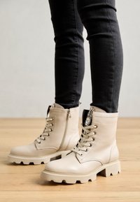 Botines de cuero beige con acabado texturizado, cierre de cordones en la parte delantera, cremallera lateral y suelas gruesas y resistentes. Combinados con jeans ajustados oscuros.