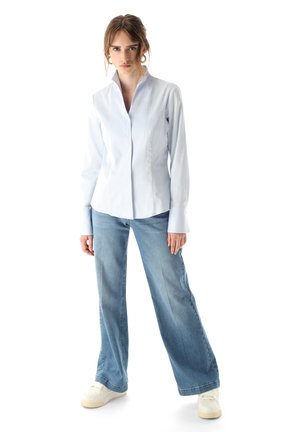 Hellblaues, eng anliegendes Hemd mit hohem Kragen, kombiniert mit weiten blauen Jeans und weißen Sneakers, alles auf einem schlichten weißen Hintergrund.