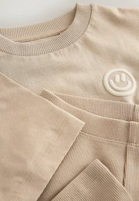 Ensemble sweat-shirt et pantalon en coton côtelé beige avec col rond et petit logo visage souriant blanc sur la poitrine du sweat-shirt.