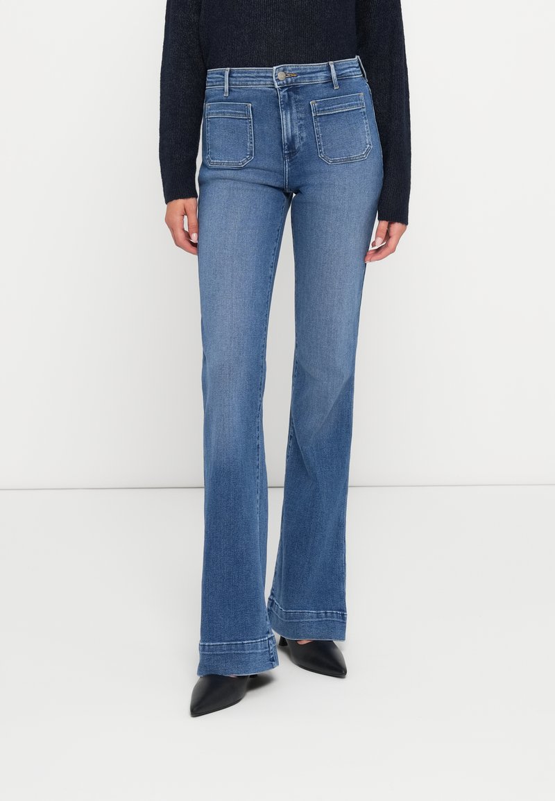 Jeans flare de denim azul com cintura alta, dois bolsos traseiros e costura em contraste. A bainha está ligeiramente desfiada para um visual casual.