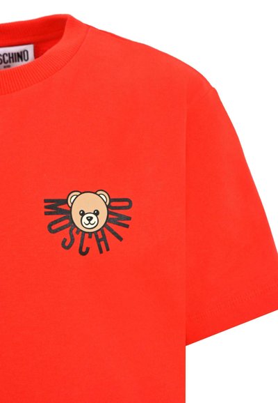 T-shirt en coton rouge avec un motif central représentant un ours en beige et noir, texte 'Moschino', encolure ronde et manches courtes.
