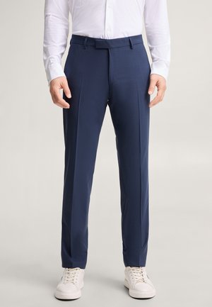 Homme portant un pantalon habillé bleu marine, une chemise blanche rentrée et des baskets blanches, debout devant un fond clair et uni.