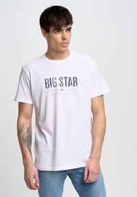 Camiseta blanca de algodón con cuello redondo, que presenta una impresión frontal en azul marino con la leyenda "BIG STAR" y "Tu Vida. Tus Jeans." en una fuente clásica.