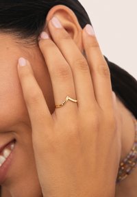 Anillo de color dorado con un diseño en forma de chevron, con un acabado suave y estilo minimalista, llevado en el dedo anular de una mano.
