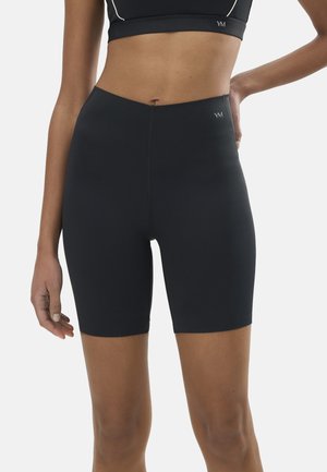 Mujer vistiendo shorts de ciclista negros de cintura alta y un top corto a juego, de pie con una mano en la cadera contra un fondo liso.