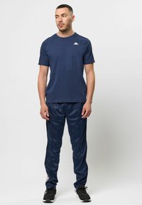 Námorně modré sportovní tričko a odpovídající sportovní kalhoty, oba elegantní a hladké, s logem na hrudi a bočními akcenty; doplněné černými teniskami.