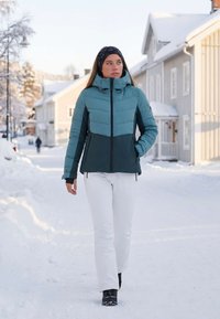 Winterjas in teal en donkergroen met padding, hoge kraag, ritssluiting aan de voorkant en twee zijzakken, gecombineerd met witte sneeuwbroek en zwarte laarzen.