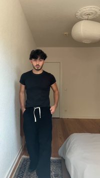 Black fitted t-shirt and loose black sweatpants with a white drawstring. Light wood floor and neutral wall background. Room features a bed and lamp.