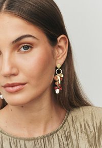 Gold-toned earrings featuring circular and teardrop shapes, embellished with red and white beads, hanging from a stud fastening.