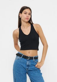 Junge Frau mit langen braunen Haaren, die ein schwarzes ärmelloses Crop-Top, blaue Jeans und einen schwarzen Gürtel trägt, steht mit einer Hand in der Tasche.