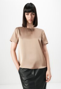 Vrouw met steil zwart haar, gekleed in een beige satijnen top met korte mouwen en een zwarte leren rok, met haar handen in haar zakken tegen een witte achtergrond.