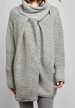 Kobieta w jasnoszarym dzianinowym płaszczu z dużym złożonym kołnierzem i wydłużonymi prążkowanymi mankietami, w połączeniu z szarymi spodniami.