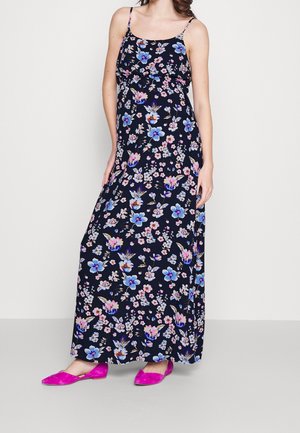Kvinde iført en sort ærmeløs maxikjole med blå og pink blomsterprint og lyserøde flade sko, stående mod en ensfarvet baggrund.