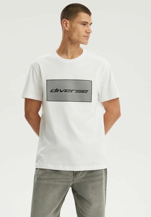 Młody mężczyzna z krótkimi włosami, ubrany w białą koszulkę z czarnym, paskowym, prostokątnym logo i szare dżinsy, stoi z rękami za plecami.