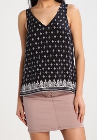 Top in cotone senza maniche di colore nero con scollatura a V, caratterizzato da motivi geometrici bianchi e un orlo inferiore dettagliato, abbinato a una gonna rosa chiaro.