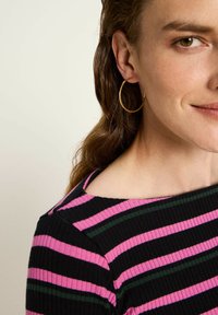 Frau mit goldenen Creolen und schwarzem, geripptem Oberteil mit rosa und grünen horizontalen Streifen, lächelnd mit sichtbarem Grübchen auf der Wange.