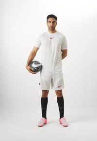 Fehér sportpóló vörös Nike logóval, fehér rövidnadrág, fekete térdig érő zoknik és rózsaszín focicipők. Fogja a fekete-fehér focilabdát.