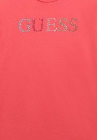 T-shirt in cotone corallo con la scritta "GUESS" impreziosita da strass multicolori in un carattere serif audace.