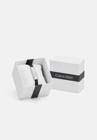 Calvin Klein karkötő fehér dobozban, fekete részletekkel. A karkötő egy elegáns ezüstszínű fém dizájnnal bír, fekete szíjjal, párnán bemutatva.