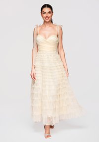 Jeune femme en robe en tulle crème à volants sans manches avec des bretelles nouées en nœuds, souriante, debout devant un fond clair uni.