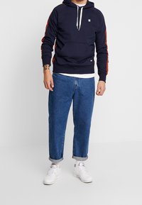 Uomo che indossa un felpa blu navy con strisce rosse, jeans blu risvoltati, calze bianche e sneakers bianche, in piedi davanti a uno sfondo bianco uniforme.